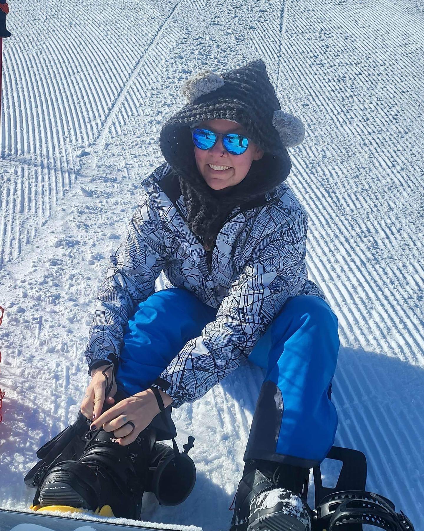Rachel in a knit hat and mirrored sunglasses, strapping into a snowboard on a snowy slope